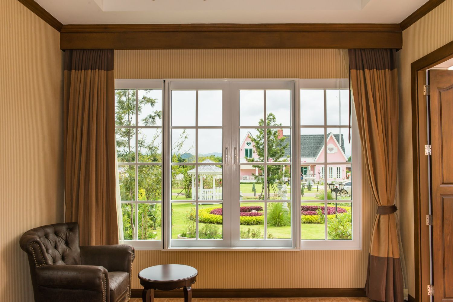 A brown leather armchair sits near a window with tan curtains, looking out onto a green yard with a small pink building.