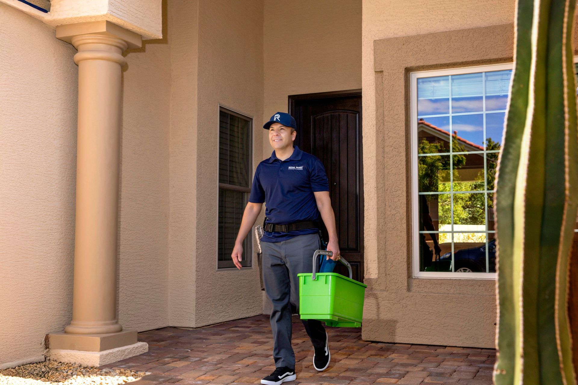 Smiling person in a blue shirt carrying a cleaning bucket, walking toward a house entrance with a window.