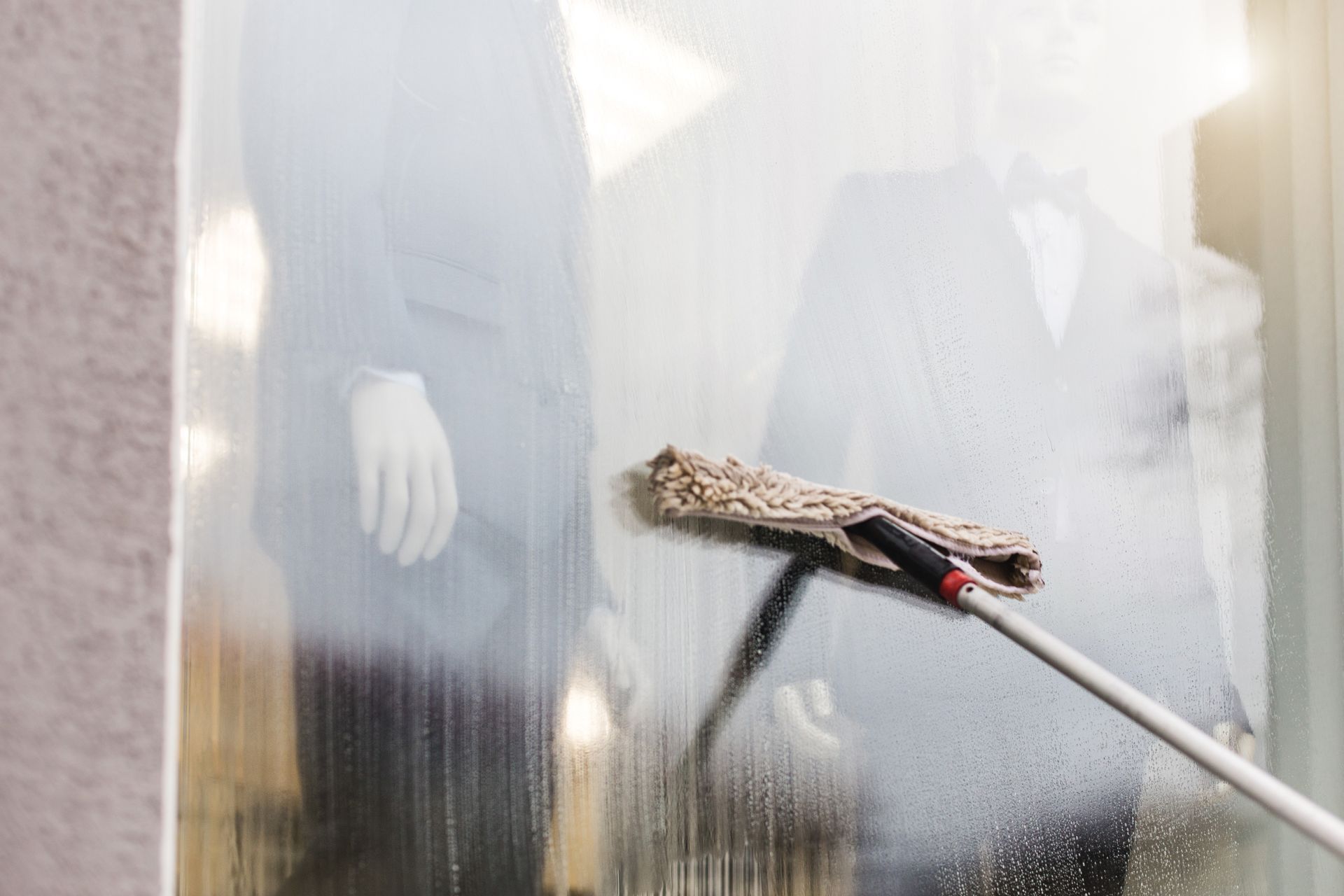 Window being cleaned with a mop, with blurry mannequins in the background.