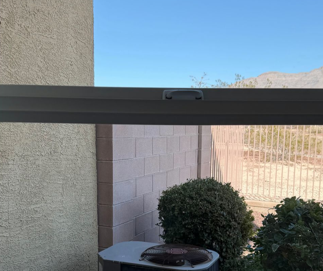 A view through a window frame showing an outdoor desert landscape with a cinder block wall, shrubs, and an AC unit.