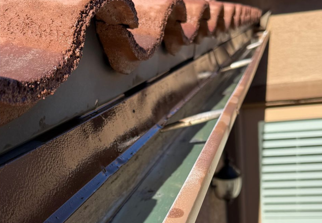 A close-up view of a metal roof gutter against terra-cotta roof tiles, showing its interior and support brackets.