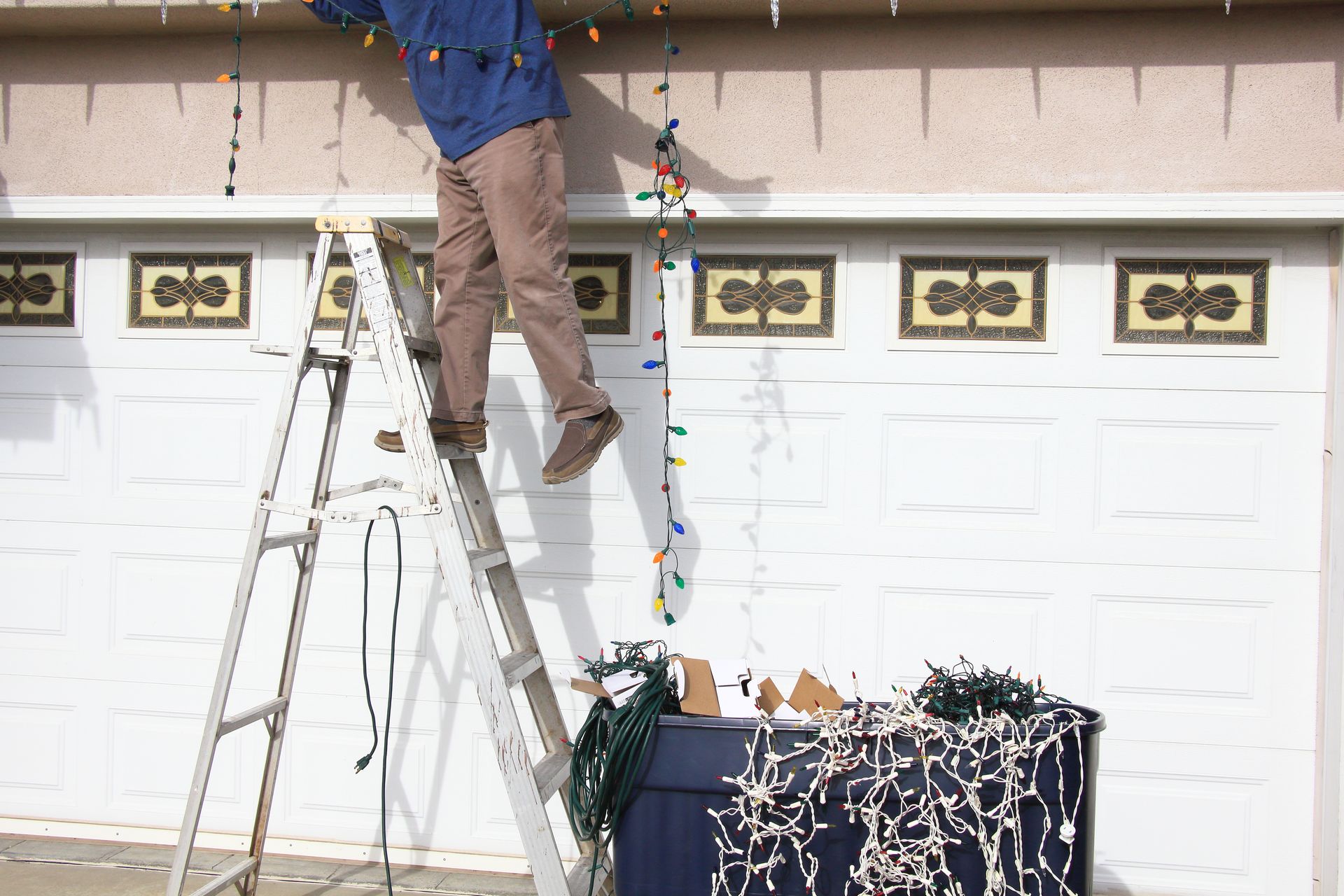 Person on ground near a ladder leaned against a house, Christmas lights strung on the roof's edge.