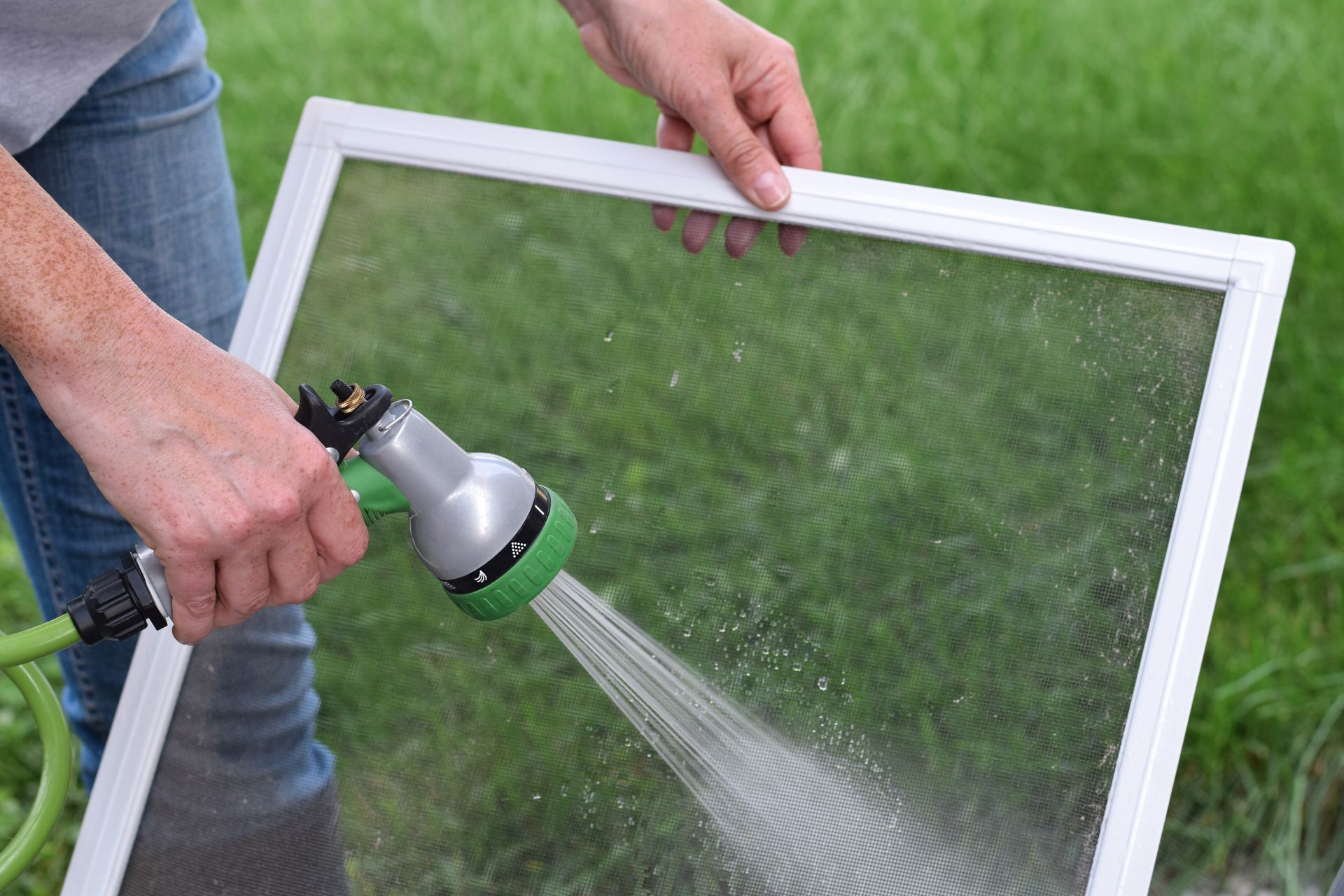 Person cleaning a white window screen with a hose outdoors.