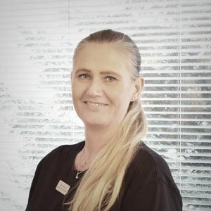 En kvinde i en sort skjorte smiler foran et vindue med persienner.