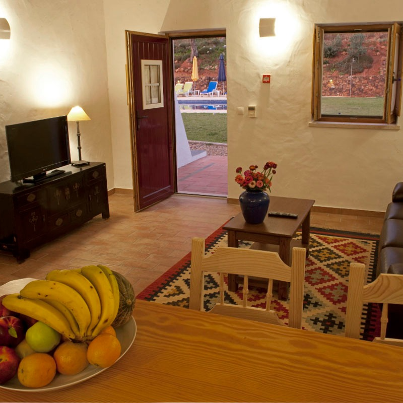 Sala de estar aconchegante com porta aberta para a piscina, fruteira, TV e pequena mesa de madeira com um vaso de flores.