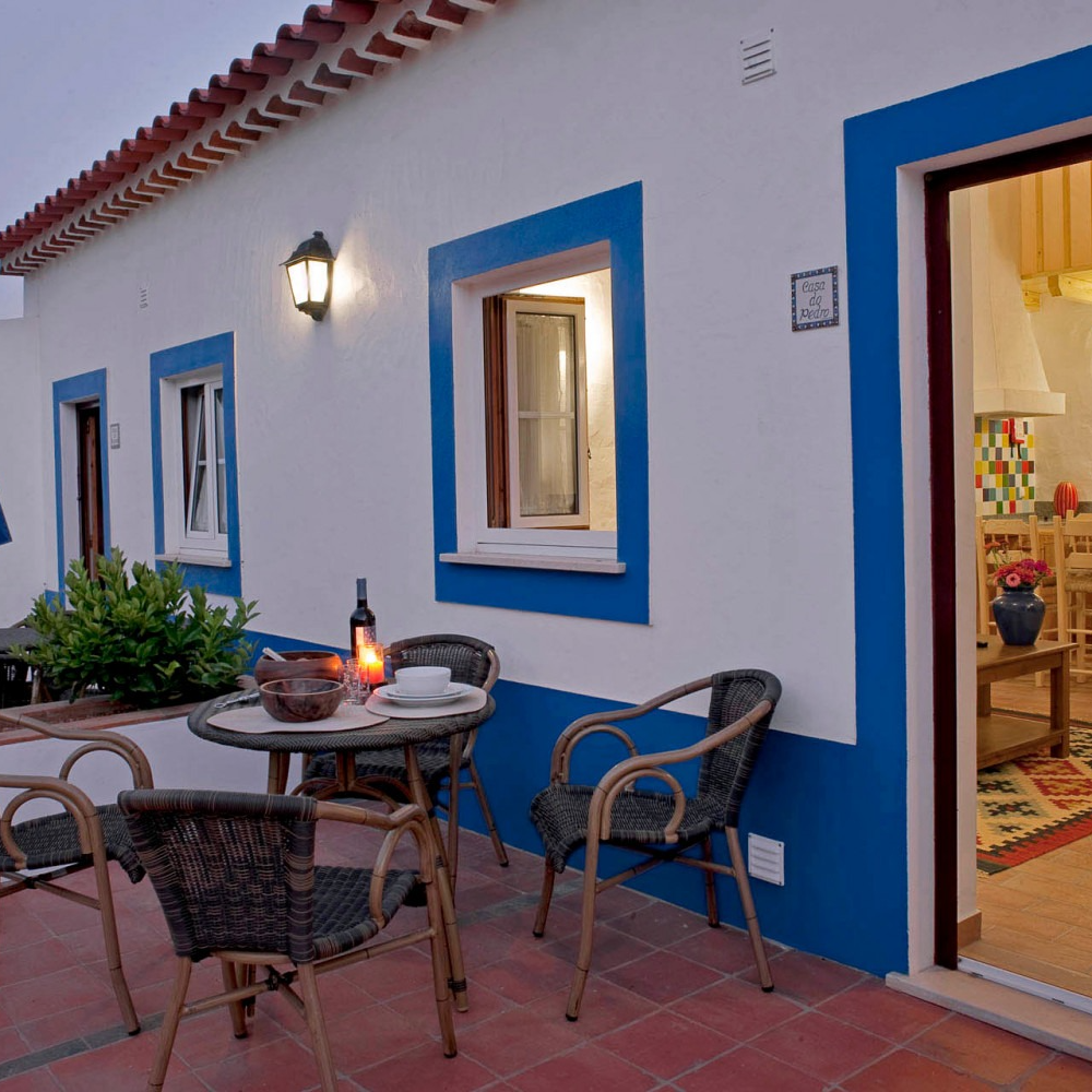 Vista exterior de um edifício branco com detalhes em azul e telhado de telha vermelha, pequeno conjunto de móveis de jardim e porta aberta.