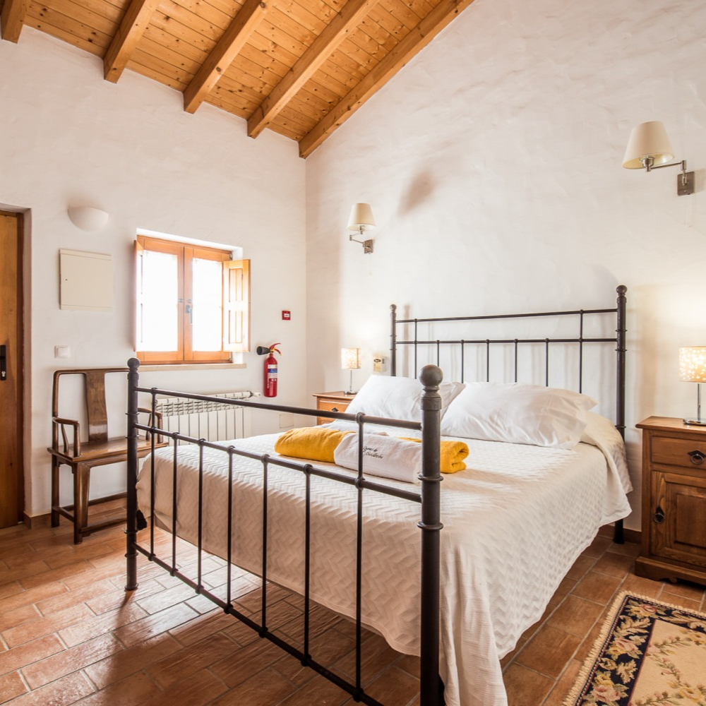 Quarto com cama de metal, teto de madeira e piso de terracota. A janela proporciona luz natural.