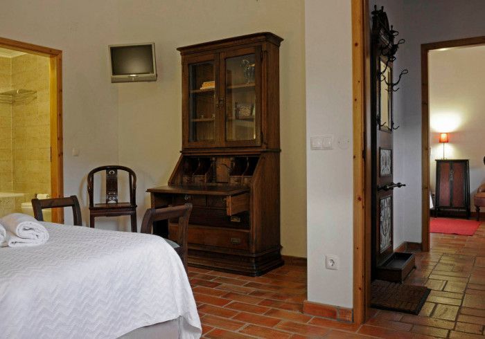 Quarto com escrivaninha de madeira, cadeiras e piso de tijolos. Um banheiro e um corredor são visíveis.