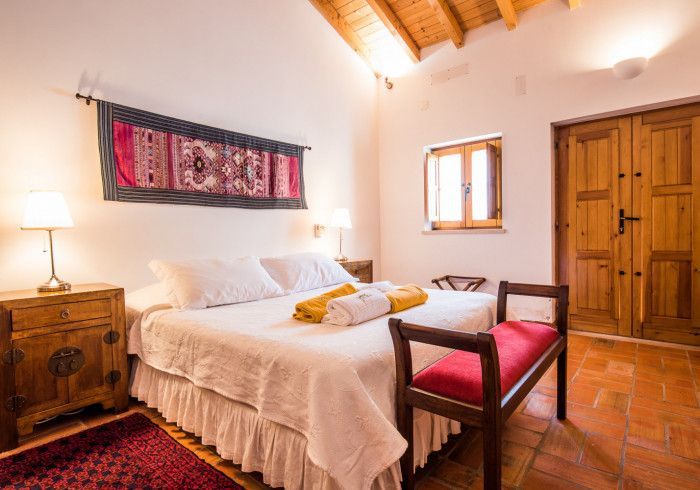 Quarto com roupa de cama branca, móveis de madeira, detalhes em vermelho e uma pequena janela.