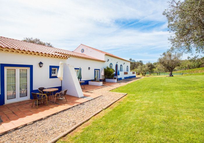 Casa branca e azul com telhado de telha vermelha, pátio externo com mesa e cadeiras, gramado verde, dia ensolarado.