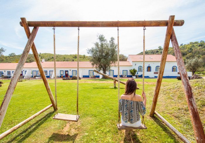 Mulher em um balanço em frente a um prédio branco com telhado vermelho, em um dia ensolarado.