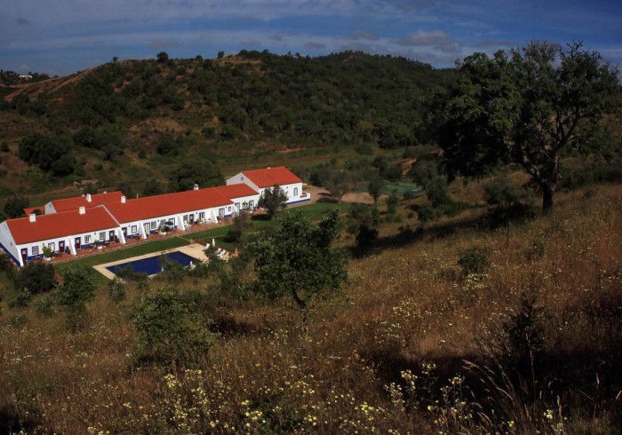 Edifícios brancos com telhados de telha vermelha e uma piscina em uma encosta com uma floresta verde-escura ao fundo.