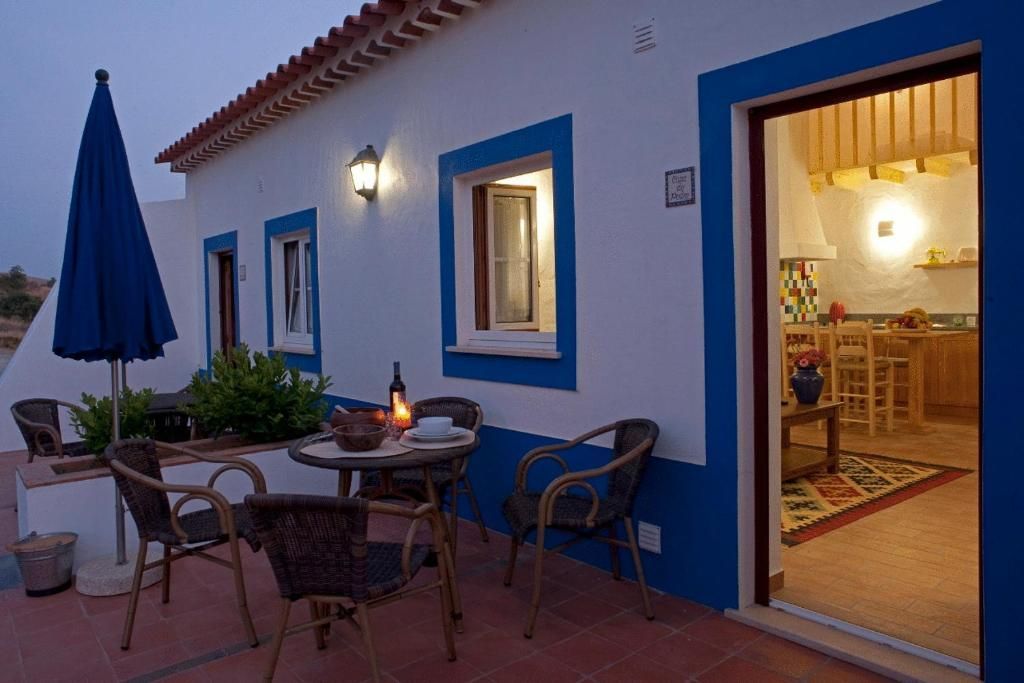 Exterior de uma casa branca e azul com uma porta aberta para uma cozinha iluminada, móveis de jardim e um guarda-sol azul.