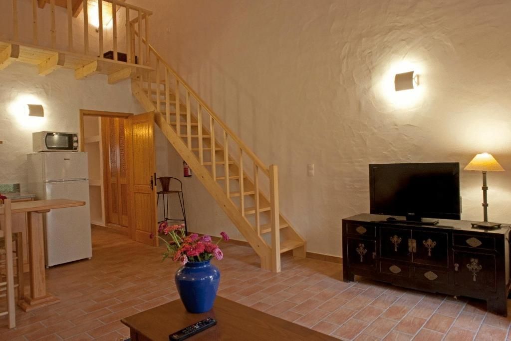 Sala de estar com escada para um mezanino. Inclui TV, armário, geladeira, micro-ondas e vaso de flores sobre a mesa.