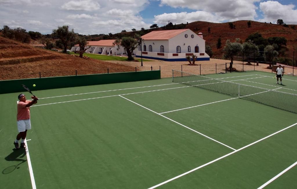 Dois homens jogando tênis em uma quadra verde em frente a um prédio branco com detalhes em vermelho, em meio a colinas onduladas.