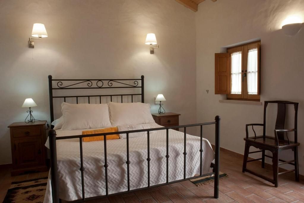 Quarto com cama de ferro forjado, mesas de cabeceira, apliques de parede, cadeira de madeira e janela com venezianas.