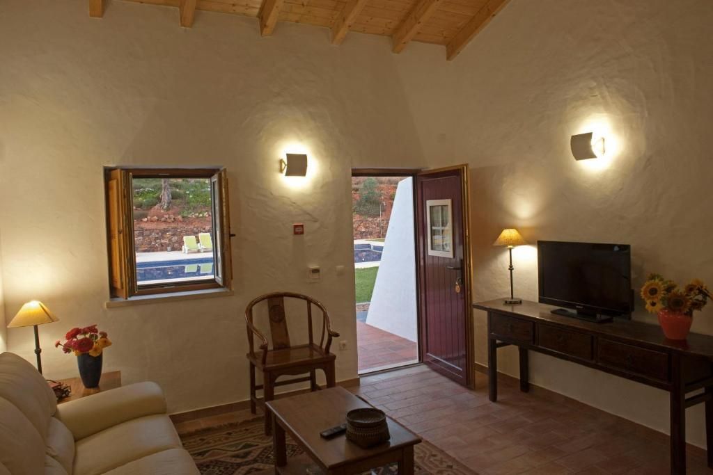 Sala de estar com sofá, poltrona, mesa, TV, janela e porta aberta. Iluminação suave e cores quentes.