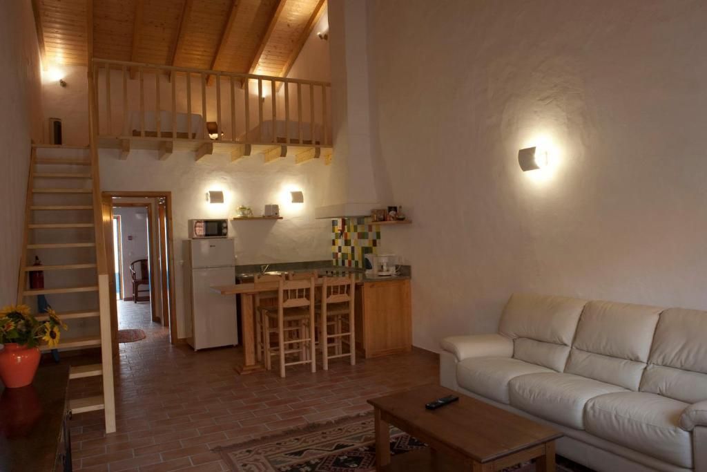 Sala de estar aconchegante com mezanino. Inclui um sofá branco, escada de madeira, cozinha e uma pequena mesa.