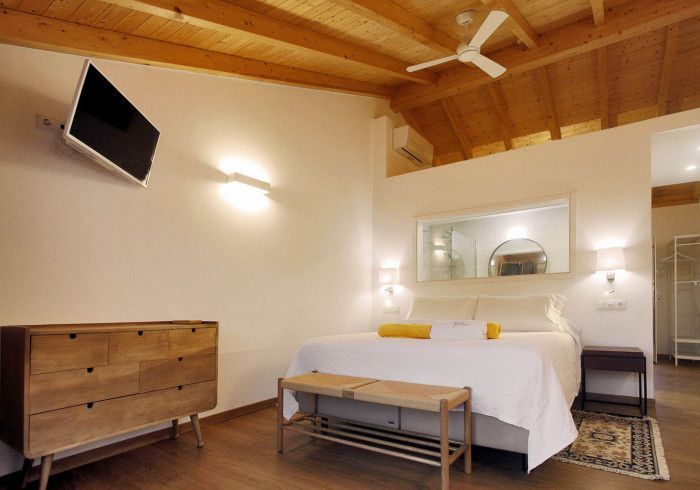 Quarto com teto de madeira, paredes brancas e uma cama grande com banco, cômoda e TV fixada na parede.