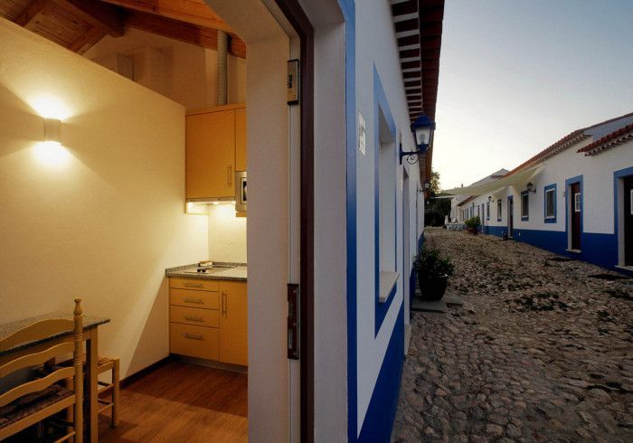 Vista de uma pequena cozinha com armários de madeira e uma fileira de prédios azuis e brancos ao longo de uma rua de paralelepípedos.