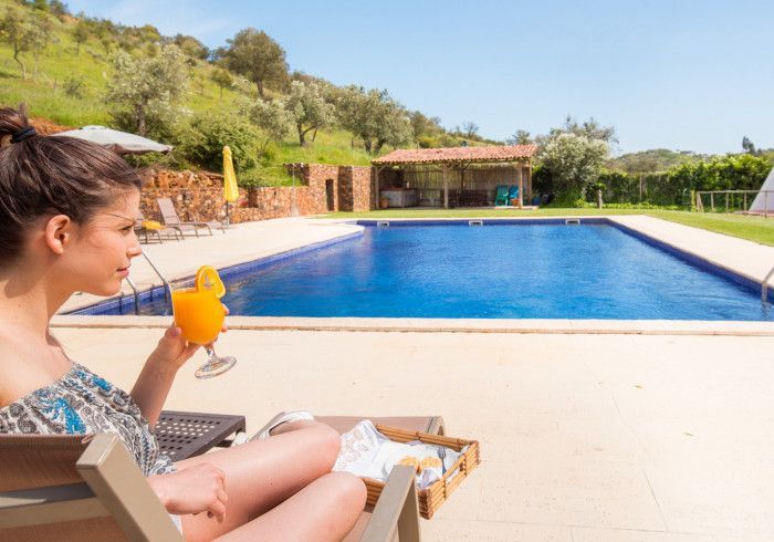 Mulher relaxando à beira da piscina, tomando um drinque, em um ambiente externo ensolarado.