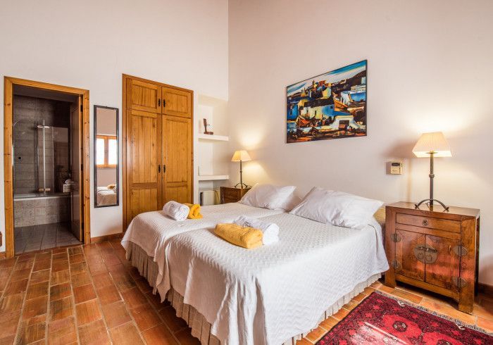 Quarto com duas camas de solteiro, móveis de madeira, piso de ladrilho vermelho, obras de arte e banheiro adjacente.