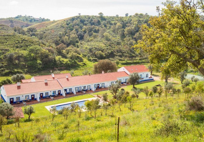 Prédio branco com telhado vermelho, piscina e árvores em uma paisagem verdejante com colinas onduladas.