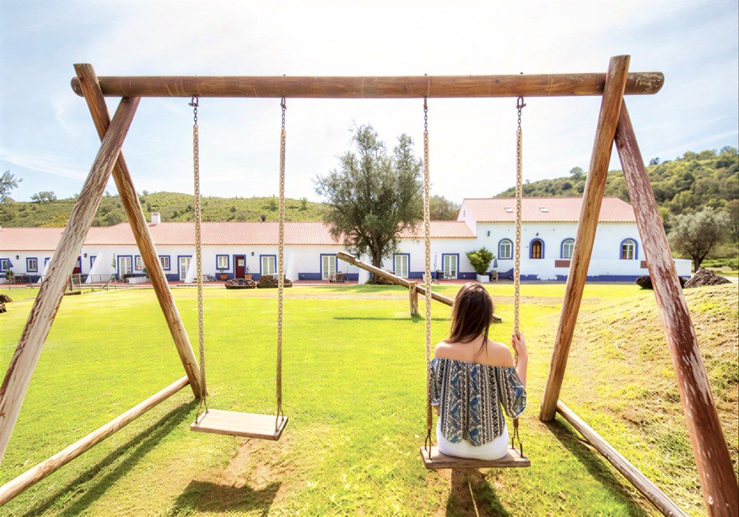 Mulher em um balanço de madeira, de frente para um prédio branco com detalhes em azul, em um campo verde.