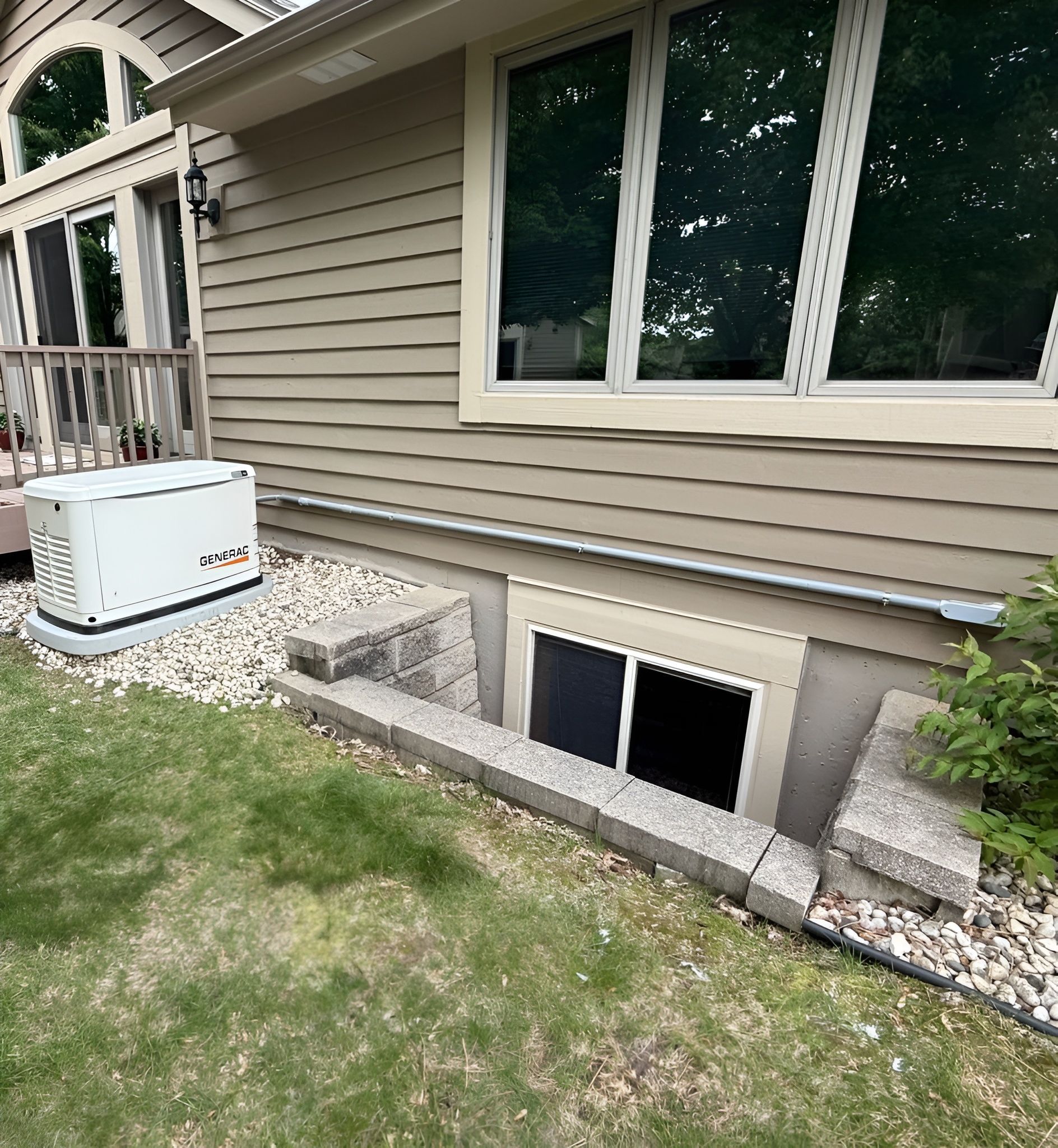 Exterior view of a house with a basement window and a generator. Beige siding, green grass, and a gray window frame.