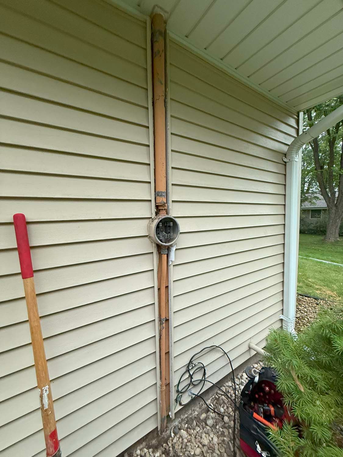 A metal pole with antennas on a beige house with a gutter.  A green bush is on the left.