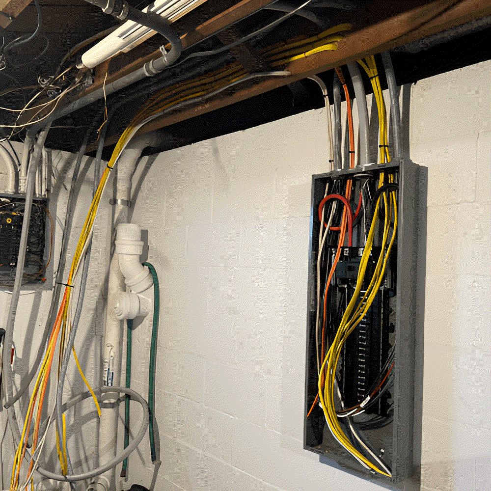 Electrical panel with exposed wires and conduit against a white cinder block wall in a basement.