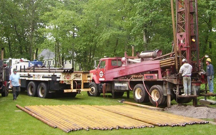 Well drilling rig on a red truck with workers, metal pipes, and a support truck on grass.