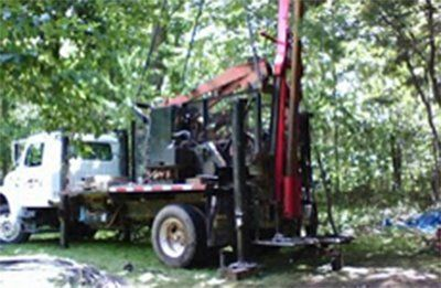 Truck-mounted well drilling rig in a wooded area, with its drilling mechanism extended upwards.