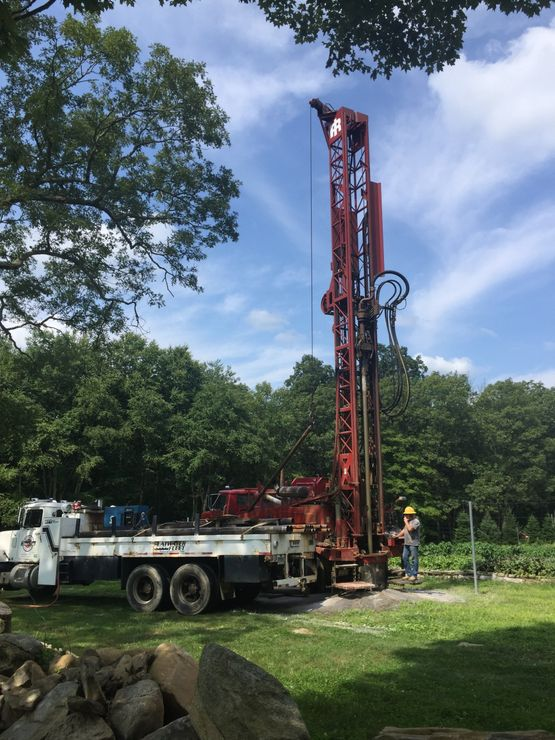 Well drilling rig on a truck, operating in a yard, with a person wearing a hard hat, sunny day.