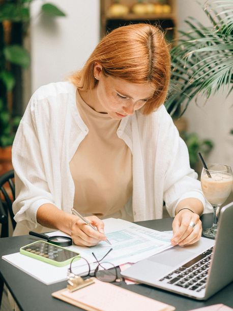 A woman sitting at a table with her laptop, glasses, a clip board, and cell phone calculator while writing on a print out.