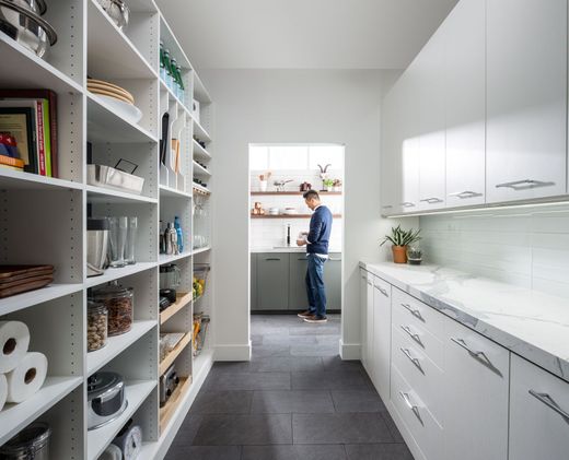Bright white kitchen with pantry shelves; a person stands at the sink near a window.