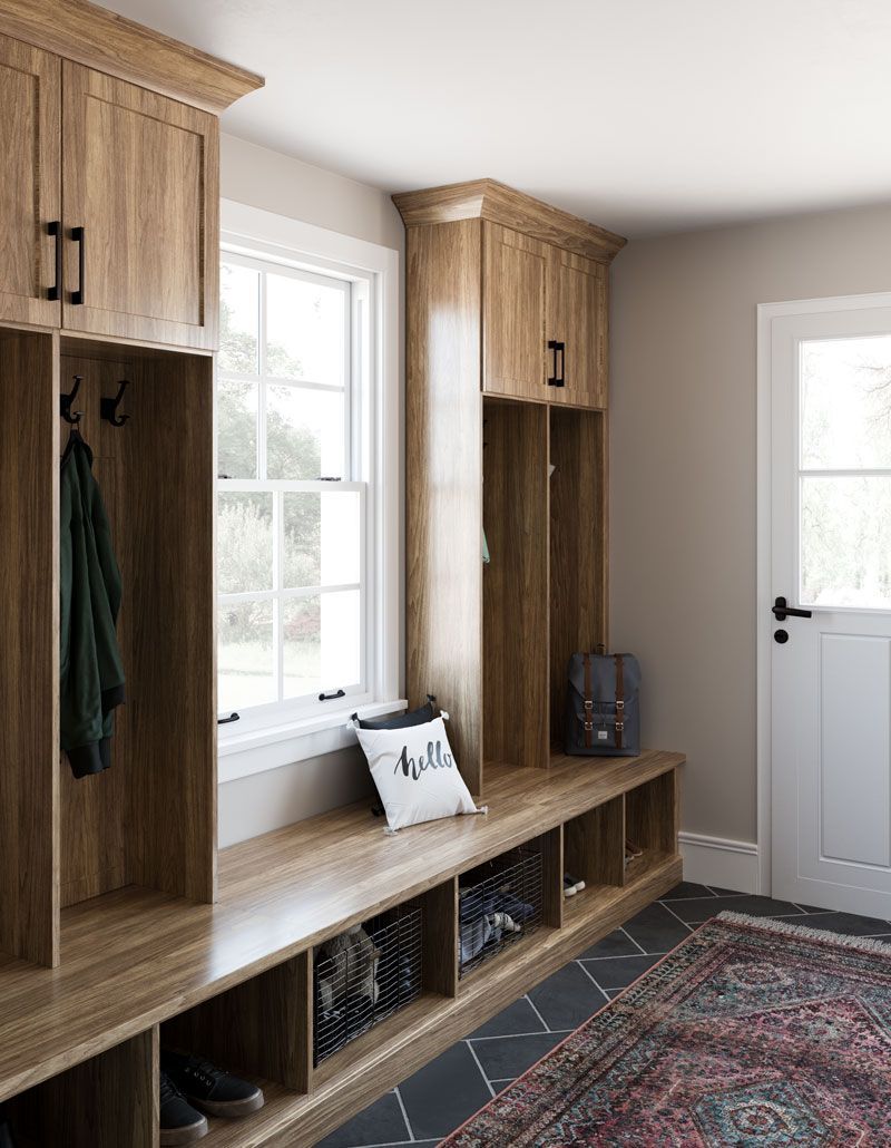 A mud room with wooden cabinets , a bench , a rug and a window.