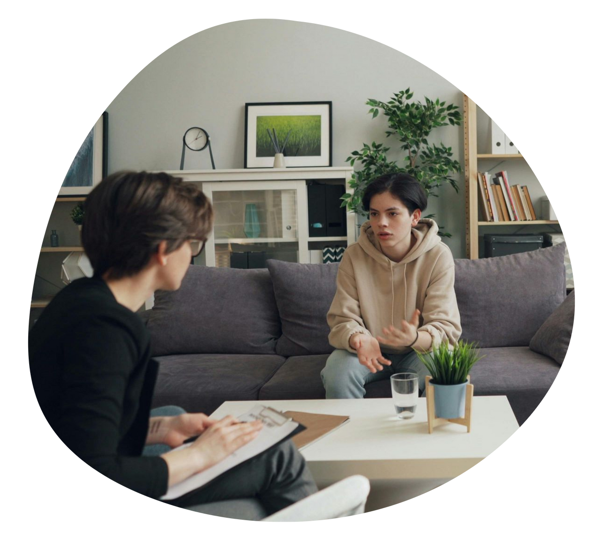 Two people talk on a sofa in a bright living room, with a coffee table, plants, and bookshelves nearby.