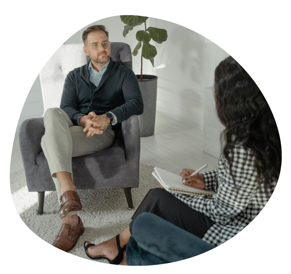 Therapist speaking with a seated client in a bright office, taking notes on a clipboard.
