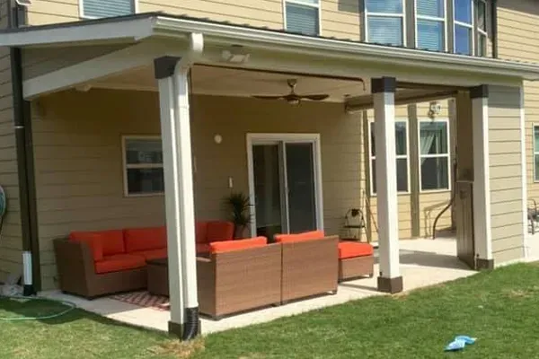 A patio with a couch and chairs under a covered porch in front of a house.