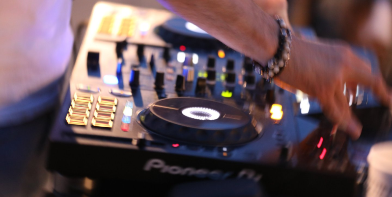 DJ che utilizza un mixer Pioneer, mani visibili, indossa bracciali e anelli, sfondo sfocato.