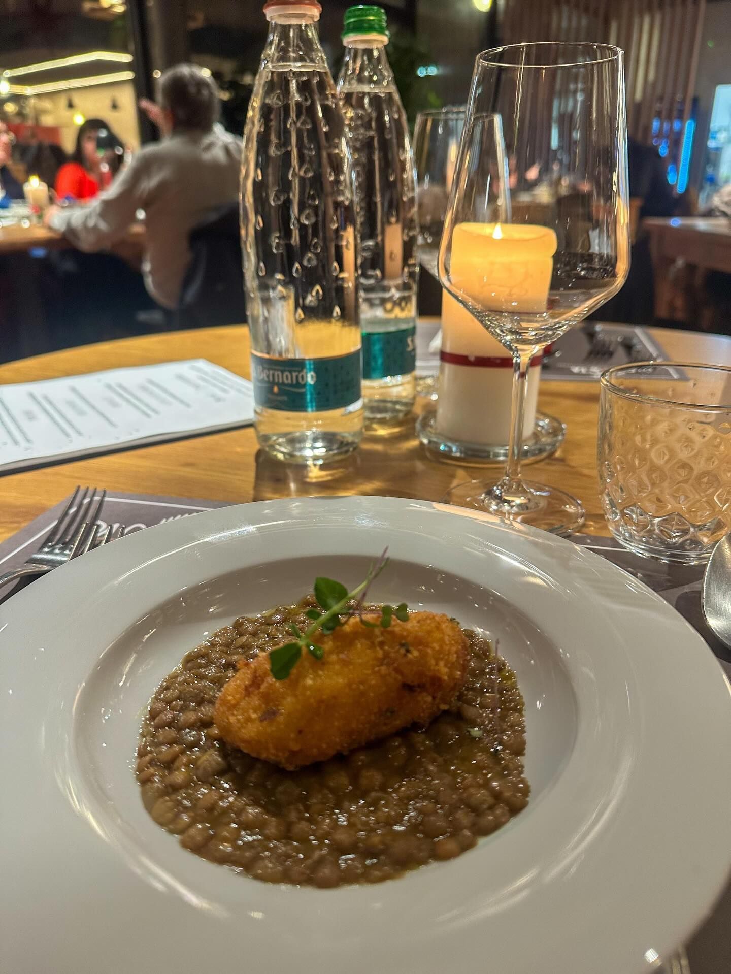 Piatto con cibo impanato sopra lenticchie, elegante ambiente da pranzo con bottiglie d'acqua e una candela accesa.
