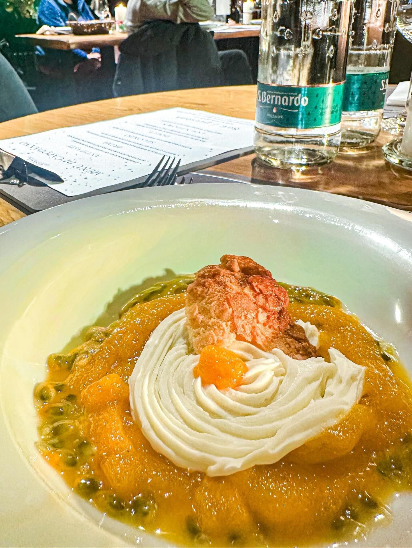 Dessert: topping bianco cremoso su frutta arancione, in una salsa gialla con semi verdi, ambientazione ristorante.