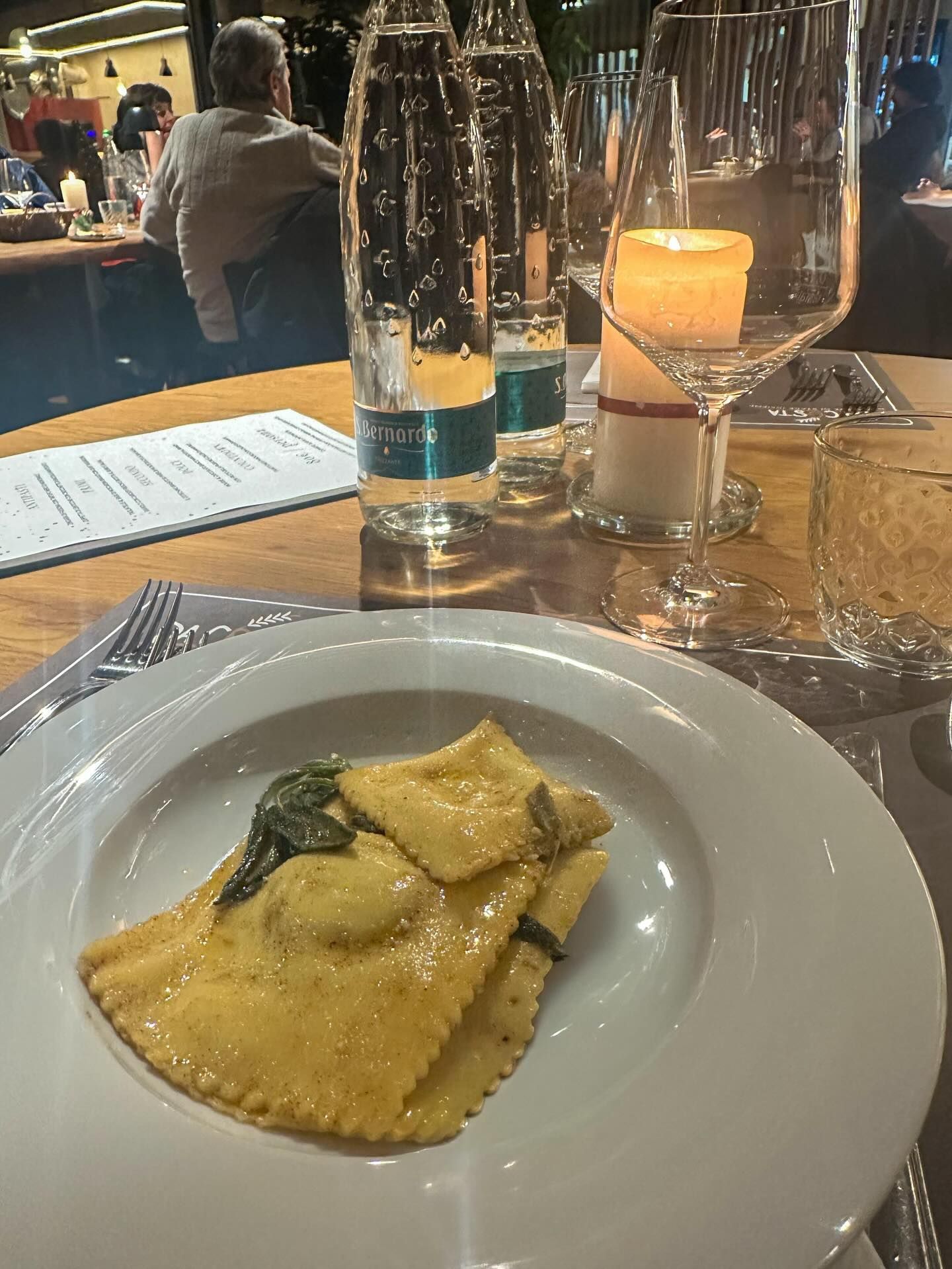Ravioli su un piatto bianco al tavolo di un ristorante. Sono visibili anche bottiglie d'acqua, bicchieri da vino e una candela accesa.