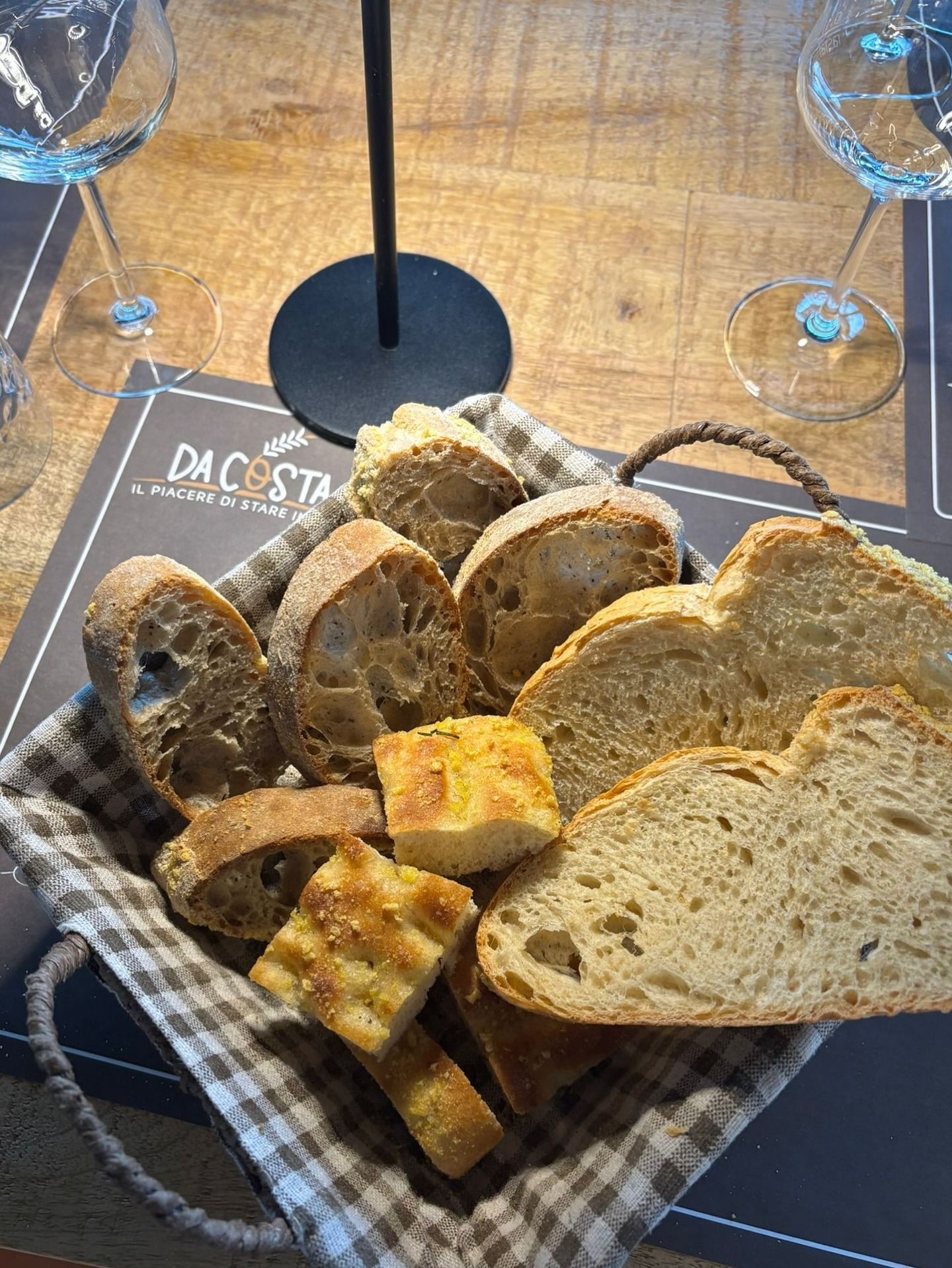 Cestino di fette di pane assortite su un tavolo di legno, accanto a due bicchieri di vino.
