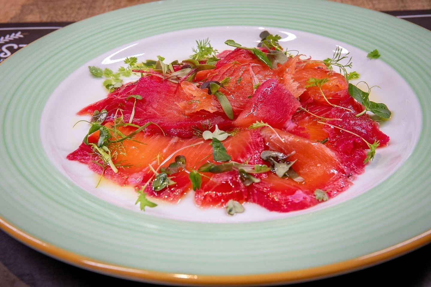 Carpaccio di salmone alle erbe aromatiche su un piatto con bordo verde. Fette di salmone rosso-arancio sono intervallate da una guarnizione verde.