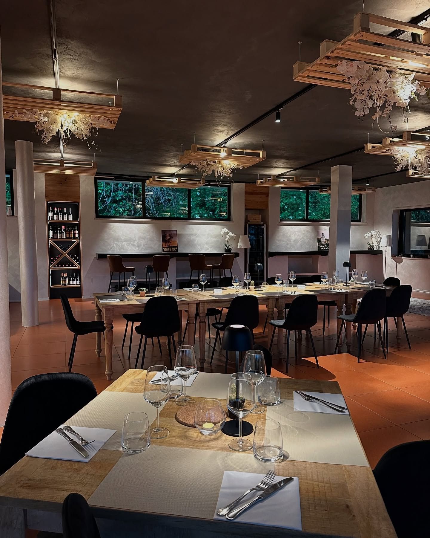 Interno del ristorante con lunghi tavoli in legno apparecchiati per cenare, sedie nere, luci soffuse e lampadari decorativi sul soffitto.