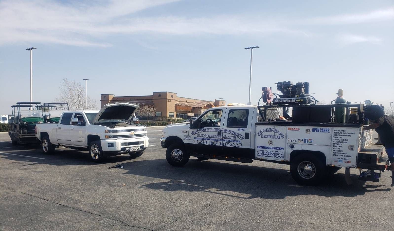 Two white pickup trucks, one with open hood and trailer, other with equipment. Bright outdoors.