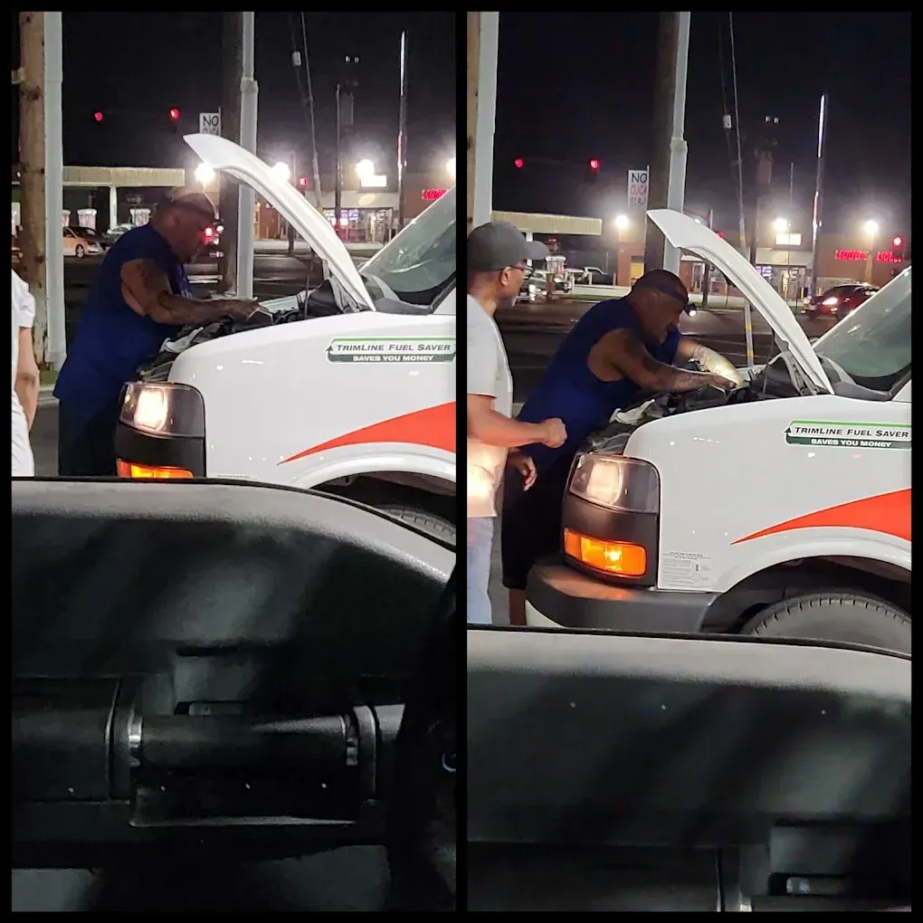 Two men working on the engine of a white U-Haul truck at night; a gas station is in the background.