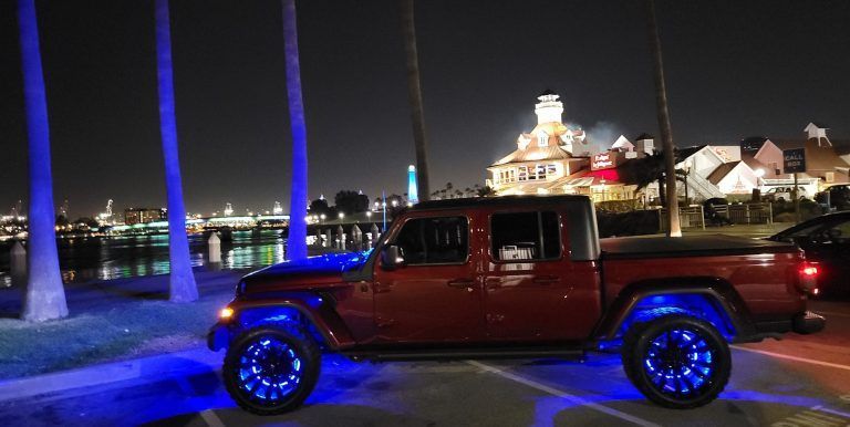 Red Jeep Gladiator with blue underglow, parked at night near water, with a lit-up building in the background.